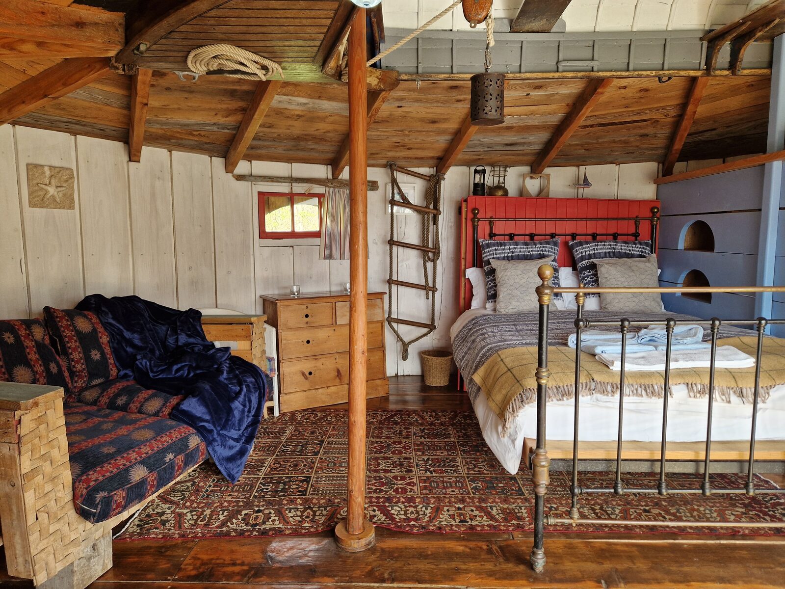 Close-up of the bed area with iron frame, red partition, rope railing, and timber ceiling