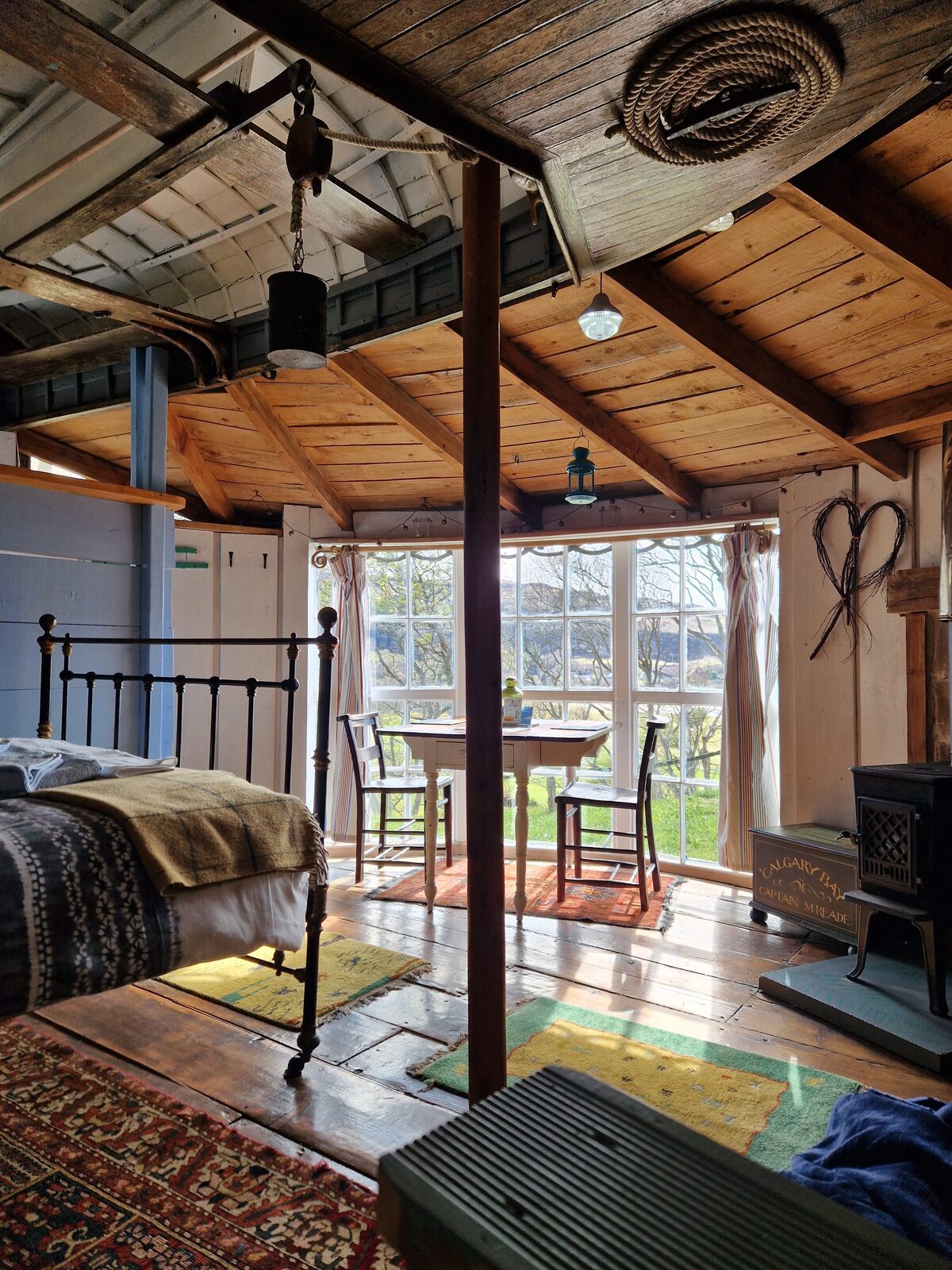 Inside Kittiwake — iron bed, dining table by curved windows, wood-burning stove, and the boat hull visible in the ceiling