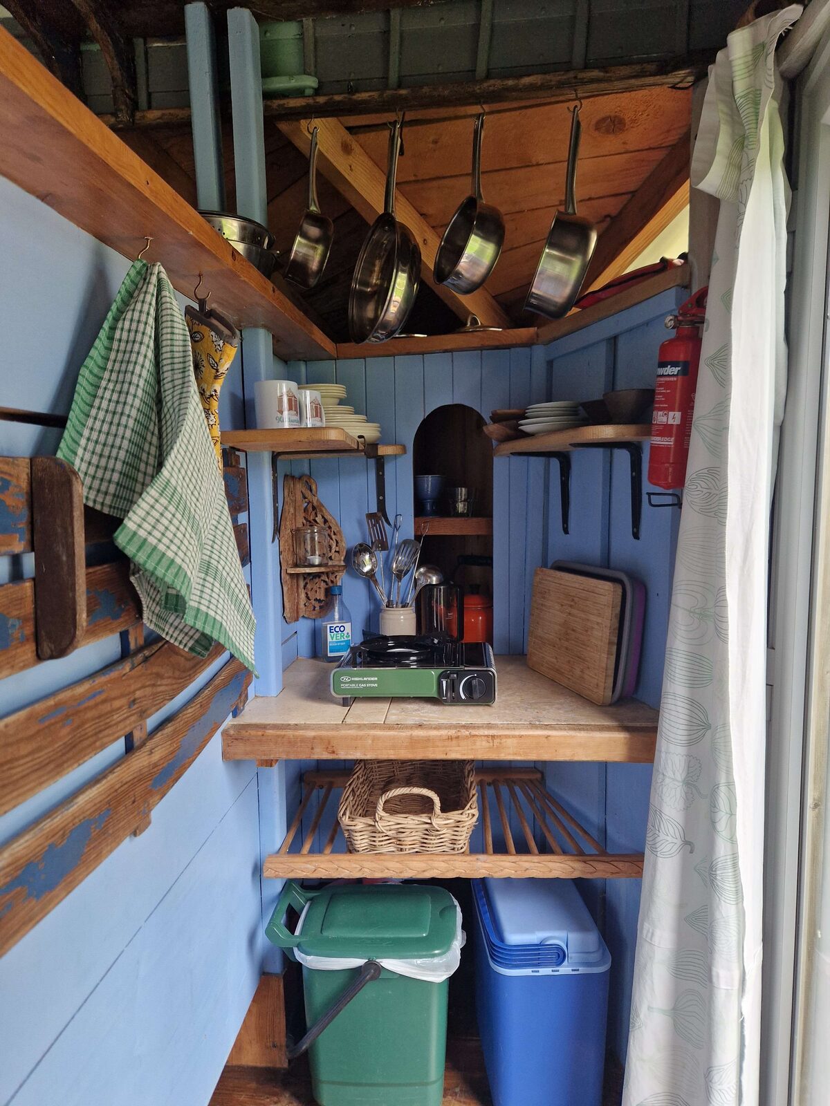 The blue-painted kitchen nook — camping stove, hanging pans, wooden shelves, and storage baskets